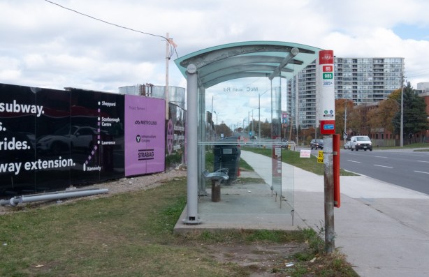 TTC bus shelter beside sidewalk on Sheppard East, hoardings or metrolinx construction site Sheppard East station, are behind the shelter