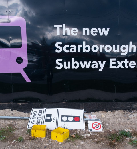 on black hoardings around metrolinx site, pink subway symbol, fallen street signs on their side at bottom of hoardings
