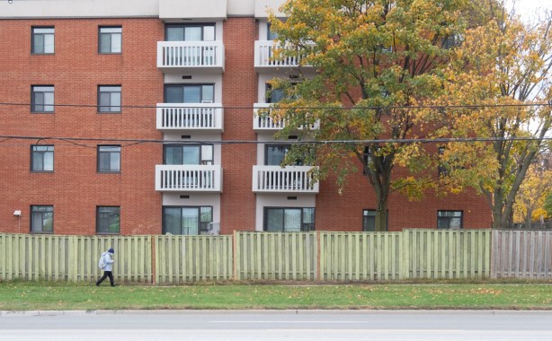 man walking past low rise apartment building with balconies, green fence beside sidewalk