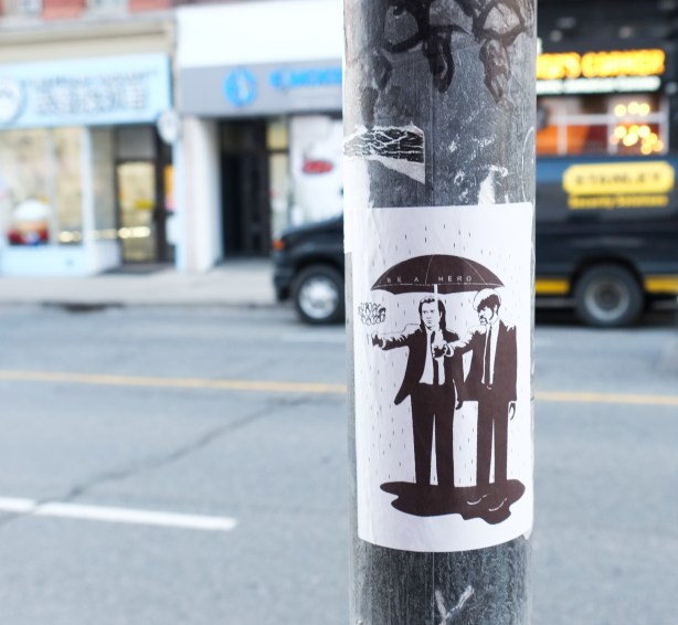 black and white slap graffiti on a pole, be a hero, a man is holding a black umbrella over the head of another men, both men in black suits