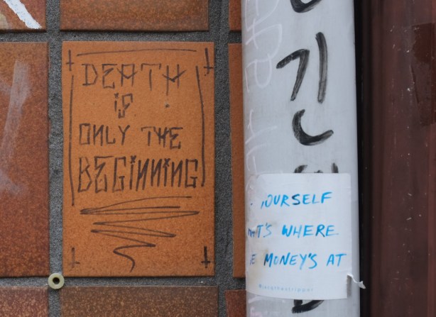 two text graffitis, small, on wall, one is written on brown tile and says death is only the beginning and the other is a white sticker written in blue