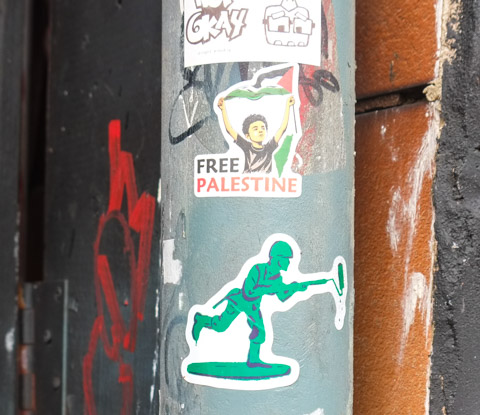 three stickers on a pole, a green army man with a paint roller in his hand, a free palestine sticker, and another