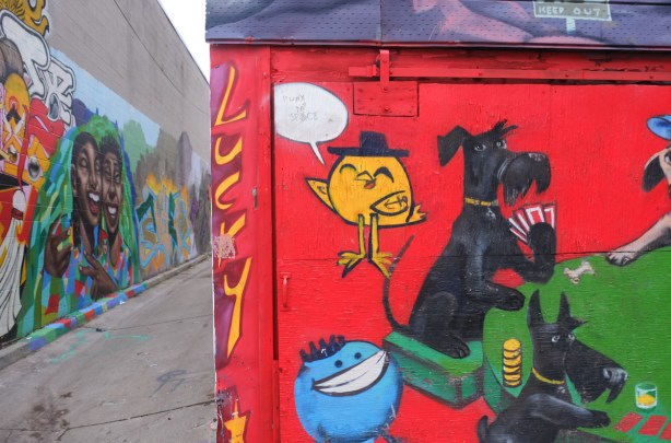 part of an uber 5000 mural of dogs playing cards, with a little yellow birdie in the top left corner, entrance to alley, other graffiti and street art can be seen 