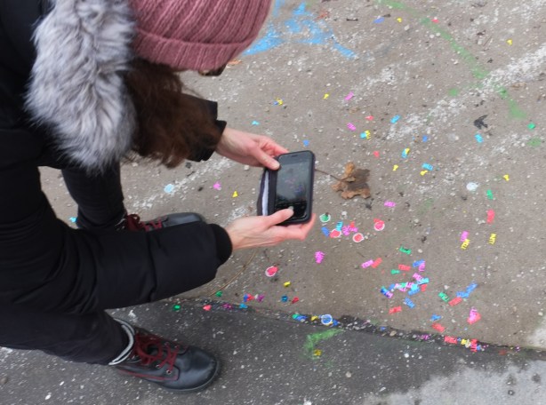 woman wearing pink hat taking a picture with her phone of confetti on the sidewalk that is bits of shiny paper spelling the word party