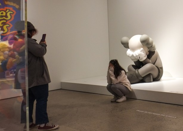 two women in an art gallery, one is taking photo of the other who is mimicking pose of sculpture's character, sitting on ground with hands over eyes