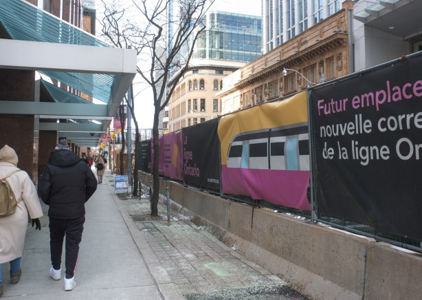 two people walking on sidewalk on Queen Street between Victoria and Yonge, fabric hoardings advertising construction of the Ontario line subway on their right