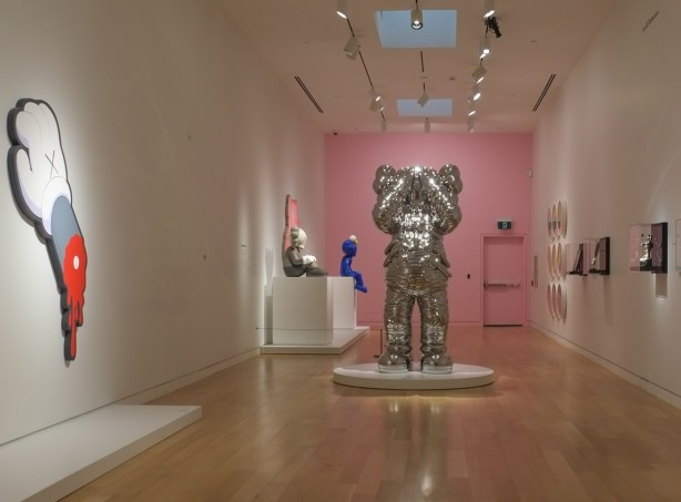 reflective silver colour KAWS family sculpture in the middle of the room in a gallery with far wall painted pink 