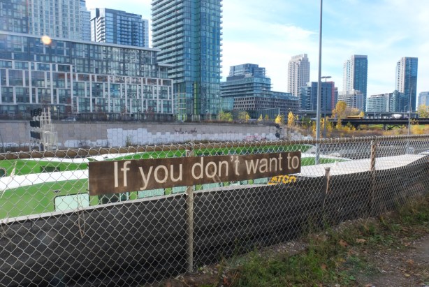 Mounted on the fence separating the railway tracks from Front Street, is a long vertical brown sign with the words if you don't want to. GO train passing behind, condo development in the background