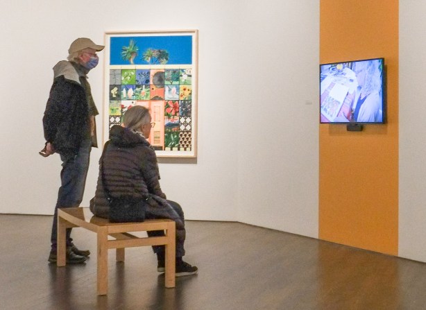 A couple at the AGO, woman is seated, man standing behind her. They are watching a video on a screen mounted on the wall 