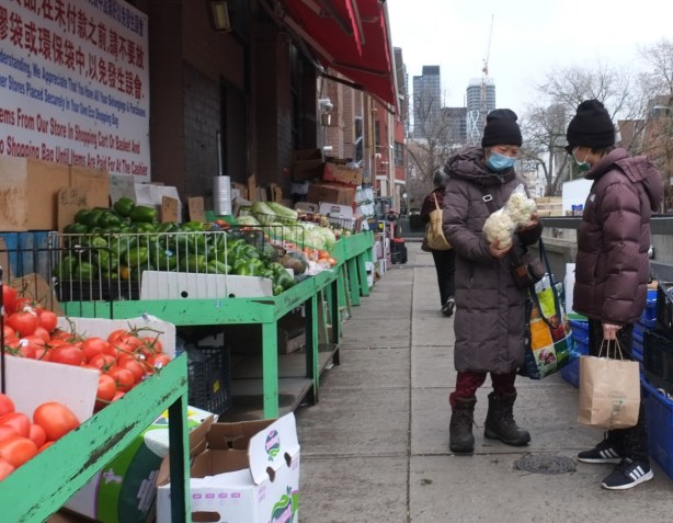 2 asian women on sidewalk, talking together, one has a clear plastic bag with 2 cauliflowers in it. 