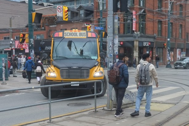 a large yellow school bus turns right onto spadina from queen street, two men are standing by the street car tracks