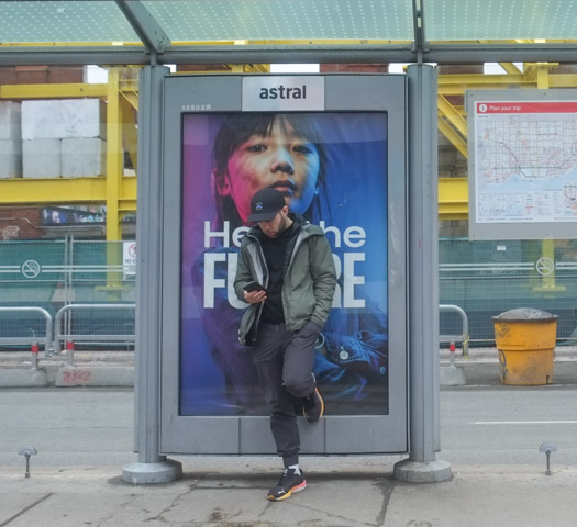 a young man is waiting for ttc streetcar on spadina, standing in front of an advertisement featuring the large face of a woman