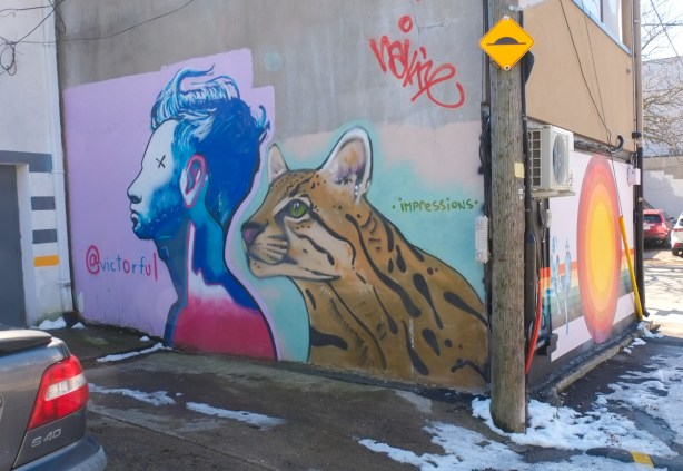 two murals in paul estrela lane, first is a man in blue with a tiger, and the other is a line of children eating ice cream while sitting beside a sun made of concentric circles, with yellow in the center and red on the outside ring 