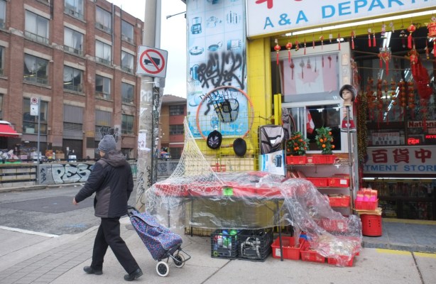 a woman walks along spadina, pulling a shopping bag on rollers, walking past a store with displays on the sidewalk