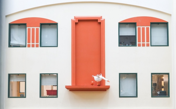 a white goose (not real) on a ledge by a fake orange door on a wall between real windows
