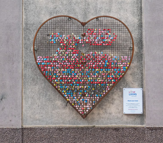 large heart shape grid of metal wires with some filled with locks, in artwork called heart locks, on wall of parking garage