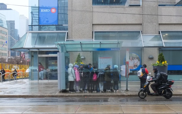 a group of people huddled together in a bus shelter on dundas, a cyclist going past them, other people on the sidewalk