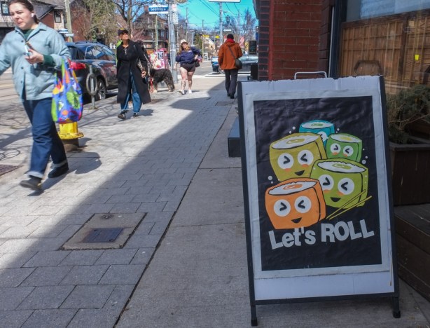people walking on sidewalk, past a sign for a sushi restaurant that says lets roll