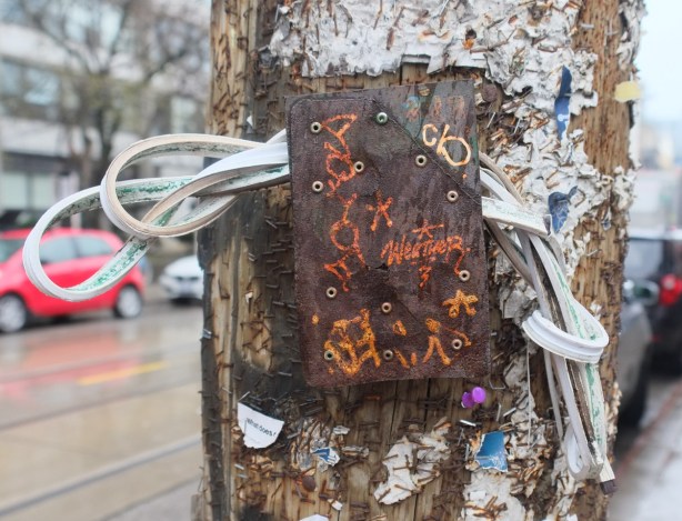 plastic coated wire bundled up and inserted between brass plaque and wood pole, lots of staples and bits of old paper in pole, artwork scratched onto the brass, including the word weather