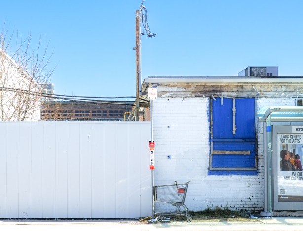 abandoned grocery store cart beside a TTC bus stop pole adjacent to white building with blue boarded over window