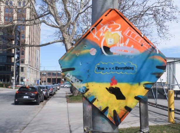 orange diamond shape construction sign that has been painted over in yellow and blue, stencil of an open hand holding a flame, text that says you are greater than everything