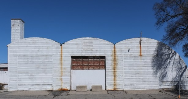 facade of white metal metal building with large garage door in center, and roofline of three curves, rusted portions,
