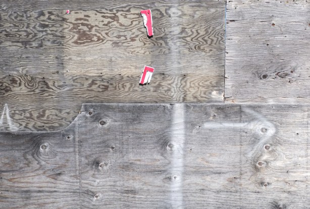 part of a wall made of weathered pieces of plywood, wood some bits of white and red paper stapled to it, a faint white arrow pointing right
