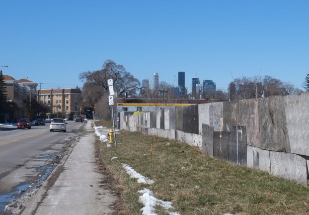 long wall of worn plywood hoardings, grass beside it, along Wicksteed, looking west towards Laird