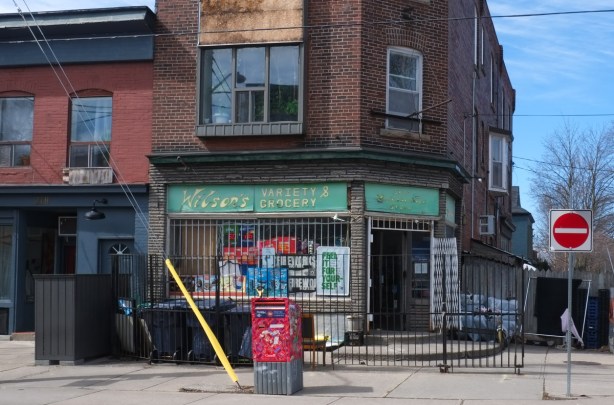 small convenience store on a corner in a residential area, Wilson's Variety and Grocery says green and yellow sign across the top of the window, black bars on the window, Canada Post mailbox in front of the store,