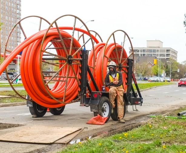 a man in hard hat and safety vest sits beside a piece of equipment used for laying large conduits for cables under roads, lots of orange conduit piping on the trailer too 
