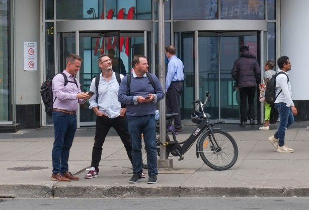 three men together on sidewalk outside Eaton Centre on Yonge Street, looking at something, one with a phone in his hand 