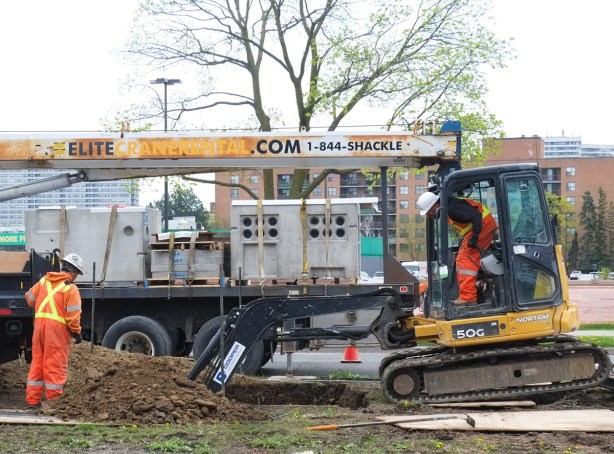 two men working beside the road, one in a digger, and one standing by the hole that it is digging