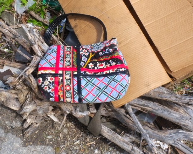 plaid handbag with ribbon trim, outside, lying on the ground, with old wood, and a flattened cardboard box