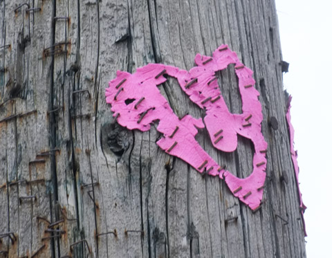 tattered pink heart with a face cut out of it, stapled to a wood utility pole that has hundreds of staples on it