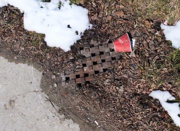 a used coffee cup lies on the ground beside a sewer drain and a small snowbank