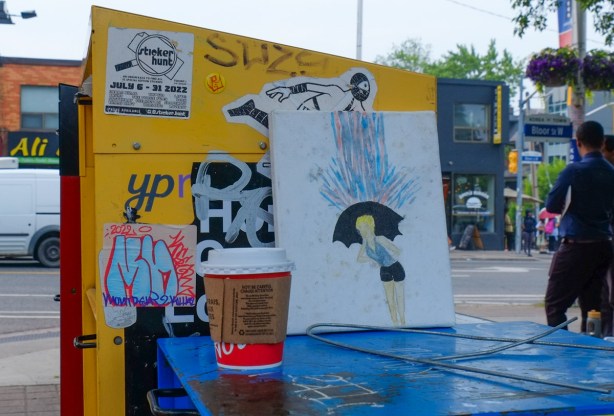 a tim hortons coffee cup on top of anewspaper box in the street, along with a painting of a woman with an umbrella in the rain. 