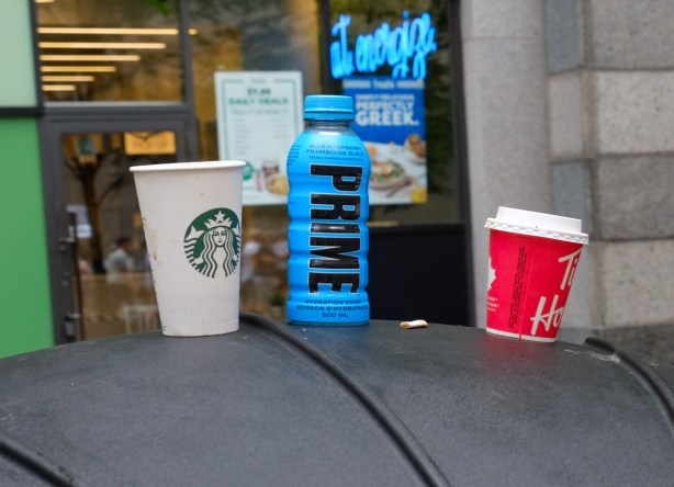 empty starbucks coffee cup, empty tim hortons coffee cup and a blue prime drink bottle, all on top of a black garbage bin on the sidewalk