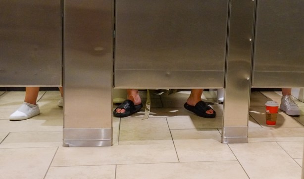 looking under the doors of bathroom stalls, three pairs of legs and feet, and one tims coffee cup