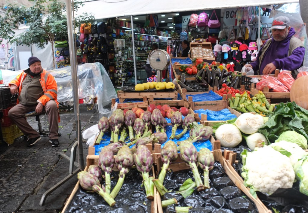 two men at an outdoor market, selling vegetables like artichokes and cauliflower, zucchini, carrots,