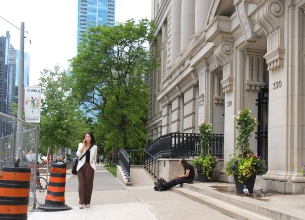 a woman on her phone walks north on university ave past the canada life building where a man is sitting on the steps