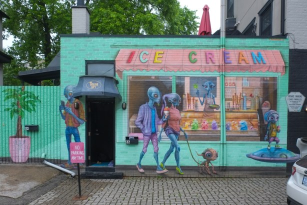 exterior of small building painted made to look like an ice cream store with big window and awning, aliens are buying and eating ice cream, also an alien dog on a leash