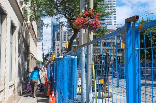 a woman carries two bags full of shopping as she walks on a sidewalk beside a blue construction fence in downtown toronto