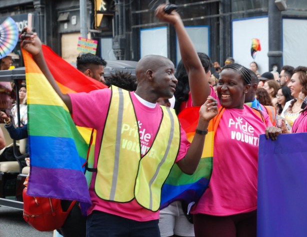 two people in pride parade, holding banner, cheering and smiling, one arm in air, wearing yellow vest, also rainbow flag over shoulders