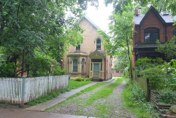 pale yellow brick parkdale house with picket fence, gravel driveway, large front yard, and old garages in the back