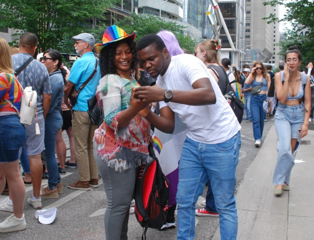 a couple looking a phone together, laughing, people behind them, pride parade