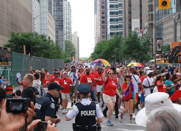 police and other people watching parade, group in red t shirts marching, as hey turn from bloor onto yonge