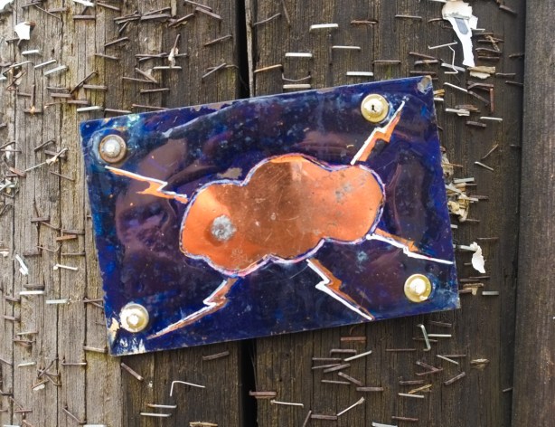 small metal street art by rocky zenyk screwed onto a wood utility pole, blue background, orange cloud and 5 streaks of lightning emanating from the cloud