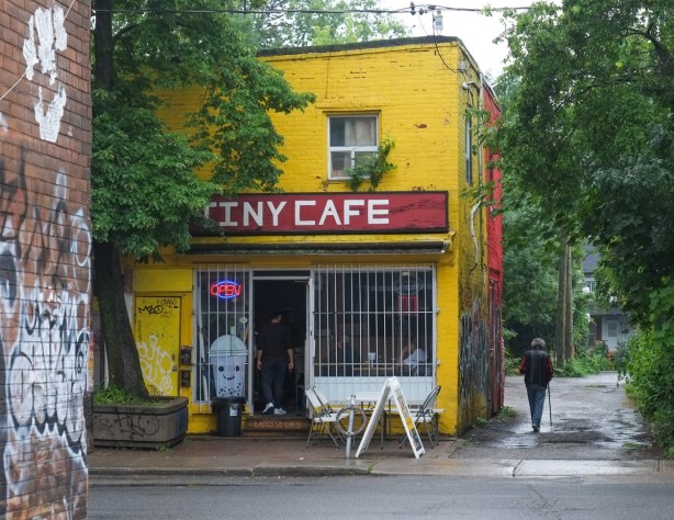 tiny cafe, a small yellow building with a red and white sign, large window in front, beside an alley in parkdale
