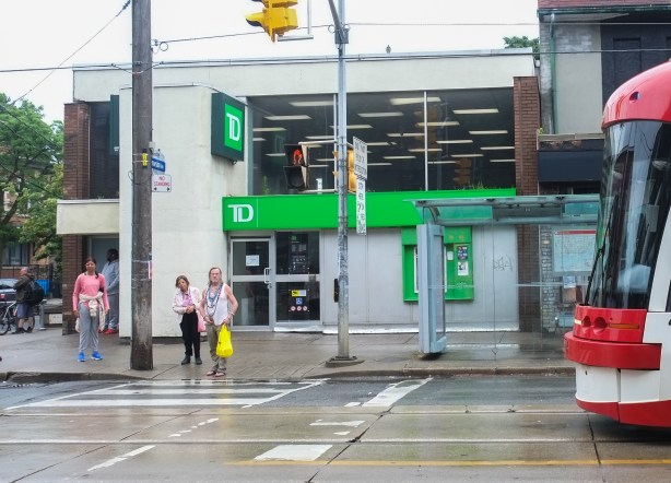 on a corner, TTC streetcar approaches a stop, with TD bank in background and people waiting for the streetcar