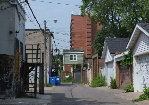 looking down a lane towards a 2 storey house with green upper storey, row of garage, midrise brick apartment building in the background
